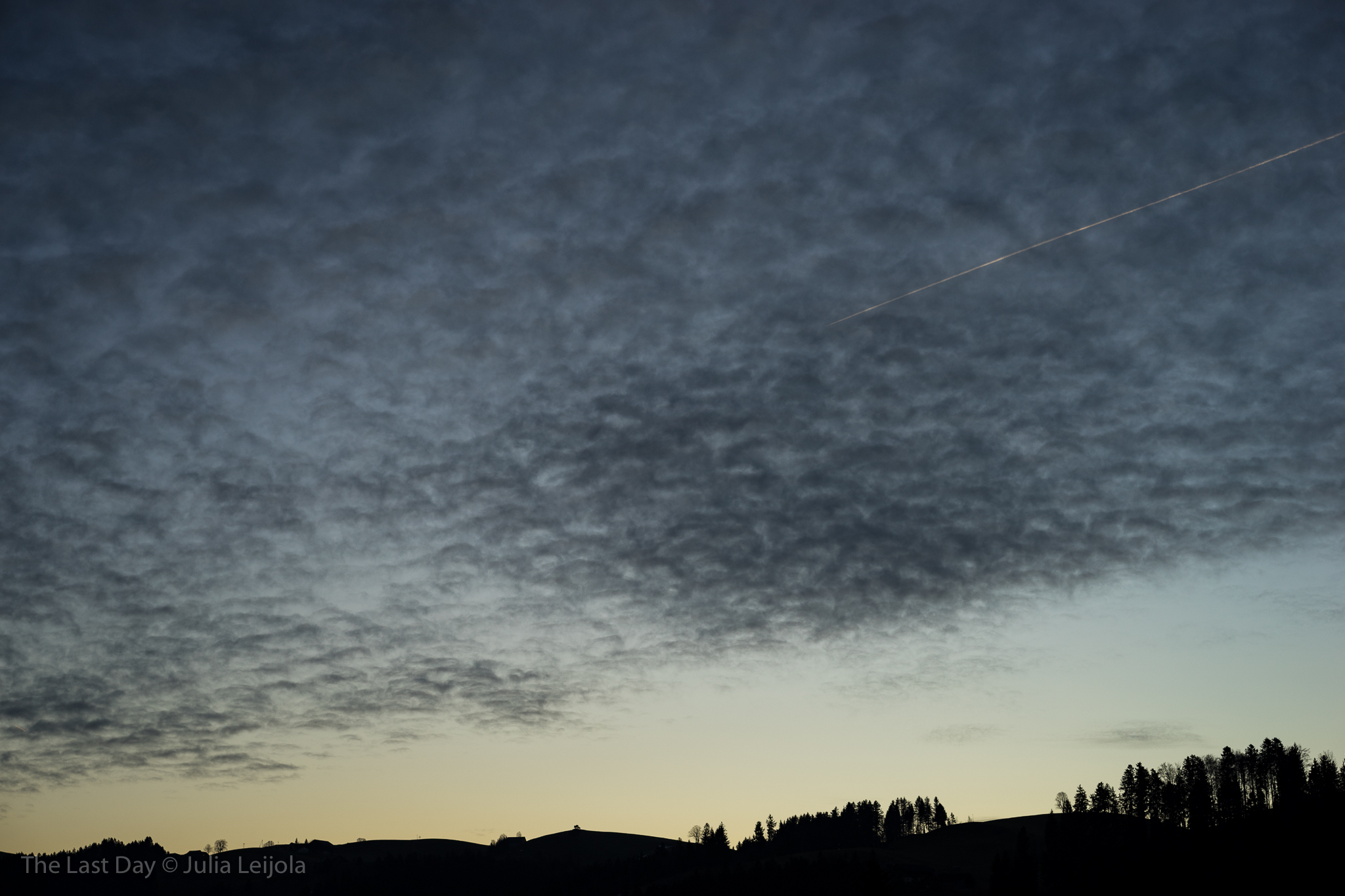 break of dawn over Dicken (SG, Switzerland): black silhouette of hills and a wide sky with a thin layer of clouds - a contrail lit up by the sun pierces the sky.